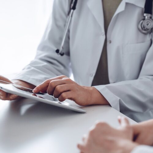 Doctor and patient sitting at the desk in clinic office. The focus is on female physician’s hands using tablet computer, close up. Perfect medical service and medicine concept